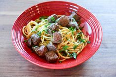 a red bowl filled with pasta and meatballs on top of a wooden table next to a fork