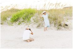 two people sitting in the sand near some tall grass and bushes, with one person taking a photo