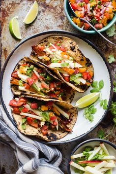 three tacos are sitting on a plate next to a bowl of salsa and lime wedges