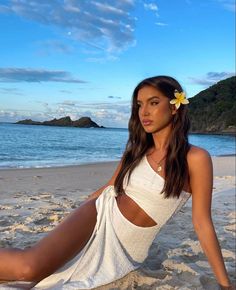 a beautiful young woman sitting on top of a sandy beach next to the ocean with a flower in her hair