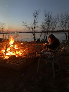 a woman sitting in front of a campfire at night with the sun going down