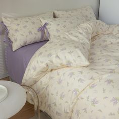 an unmade bed with purple and white flowers on the comforter next to a night stand