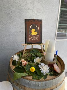 a wooden sign sitting on top of a barrel filled with flowers and leaves next to a candle