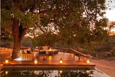 an outdoor deck with candles on it next to a pool and trees in the background