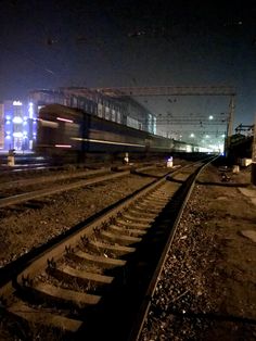 the train is going down the railroad tracks at night, with its lights on and buildings in the background