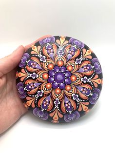 a hand holding a painted rock with an orange and purple flower design on the top