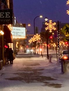 people walking down a snowy street at night with christmas lights on the buildings and signs