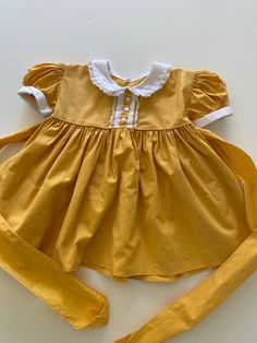 Vintage 1950's era little baby dress.  Golden yellow cotton with white sleeve cuffs, peter pan collar, decorative buttons and lace accents.  Full gathered skirt. Buttons and ties in the back.  Excellent vintage condition.   label:  none; looks to be handmade fabric:  cotton size:  none; size 6-9 months is just an estimate.  Please check measurements for an accurate fit.  Questions are welcome! dress was measured across the front side lying flat underarm to underarm 10 1/2" length 15" shoulder 9" Vintage Cotton Dress For Dress-up, Classic Cotton Dress With Lace Collar, Cotton Dress With Peter Pan Collar And Buttons, Fitted Cotton Vintage Dress With Doll Collar, Yellow Puff Sleeve Cotton Dress, Yellow Cotton Puff Sleeve Dress, Yellow Cotton Retro Vintage Dress, Vintage Cotton Dress For Dress-up Occasions, Vintage Doll Collar Dress For Dress-up