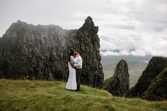 a man and woman standing on top of a lush green hillside next to tall mountains