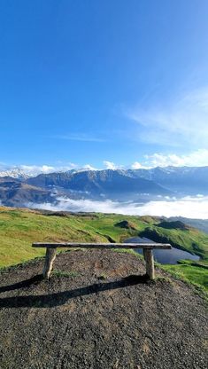 a wooden bench sitting on top of a dirt hill next to a green field with mountains in the background