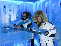 two people standing in an ice hotel with snow equipment on their hands and one person wearing a fur hat