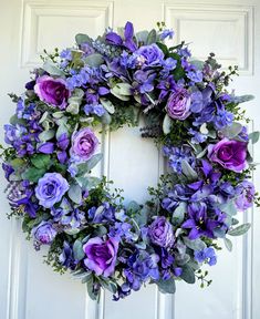 a wreath with purple flowers and greenery hanging on a white front door in the daytime