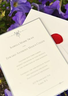 a wedding card with a red button on it next to purple flowers