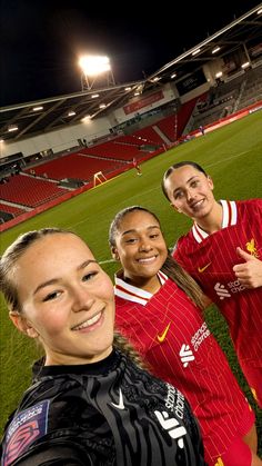 three women are posing for a photo in front of an empty soccer field at night