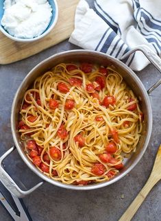 a pan filled with spaghetti and tomatoes on top of a table next to a wooden spoon