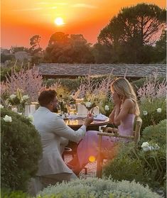 a man and woman sitting at a table in the middle of a garden with sunset behind them