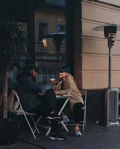 two people sitting at a table in front of a store window talking and drinking coffee