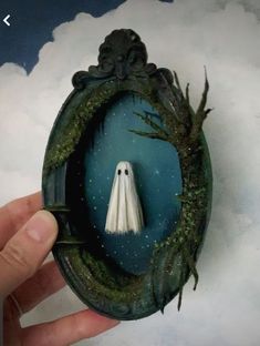 a hand holding a miniature white ghost in a frame with trees and clouds behind it