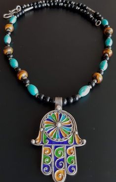 Handmade vibrant Tuareg Sterling silver and enamel 'Hamsa' necklace. 5 cm high Hand of Fatima centerpiece is decorated with intricate silver filigree details which border the traditional blue, green, turquoise and gold enamel (made using fire to fuse special glass powder onto the silver). The necklace itself consists of reconstituted turquoise beads, pretty camel bone beads, black glass and metal beads, finished with a pretty silver closure. *PLEASE NOTE THAT YOUR PHONE NUMBER IS REQUIRED SEPARATELY TO INITIATE SHIPPING* Blue Enamel Bohemian Necklaces, Bohemian Silver Enamel Necklaces, Traditional Blue Necklaces With Inlay, Traditional Blue Filigree Jewelry, Traditional Blue Necklace With Inlay, Traditional Multicolor Necklace With Inlay, Traditional Blue Inlay Necklaces, Traditional Sterling Silver Inlay Necklaces, Silver Amulet Necklace With Inlay