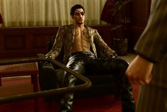 a man sitting in a chair with no shirt on, wearing black leather pants and a gold jacket