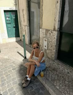 a woman sitting on the side of a building with her legs crossed and wearing sunglasses