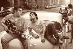 two men sit on the hood of a car while another man plays an acoustic guitar