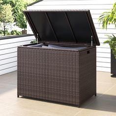 an outdoor wicker storage box with lid open on the ground next to a potted plant