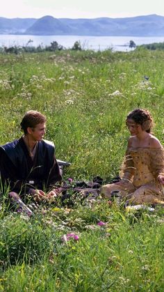 a man and woman sitting on the ground in tall grass with mountains in the background