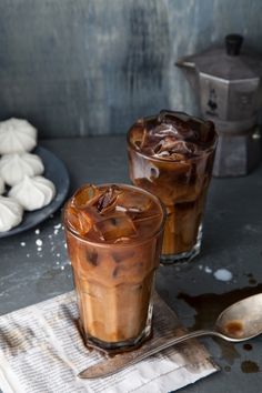 two glasses filled with iced coffee sitting on top of a table next to spoons