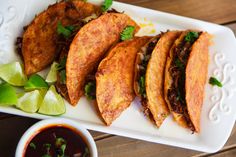 some tacos are sitting on a white plate next to dipping sauce and lime wedges