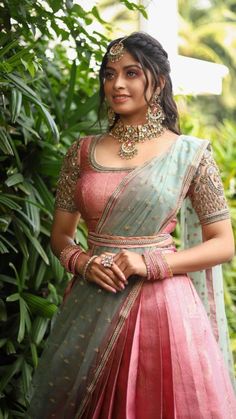 a woman in a pink and green lehenga standing next to some trees with her hands on her hips