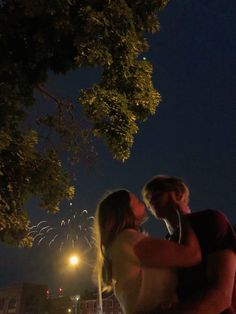 a man and woman standing under a tree at night with fireworks in the sky behind them