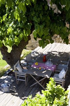 a table and chairs on a wooden deck under a tree with water in the background