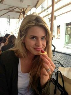 a woman sitting at an outdoor table with food in her hand and looking into the camera