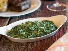 spinach in a white dish on a wooden table with wine glasses and plates behind it