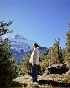 a woman standing on top of a grass covered hillside next to trees and mountains in the background