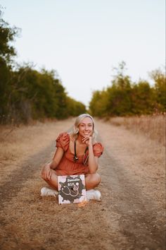 a woman sitting on the side of a dirt road holding a chalkboard with a sign in front of her