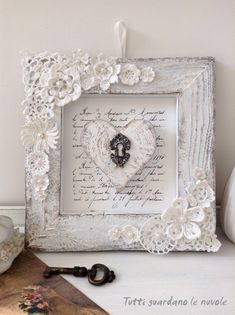 a white frame with an ornate heart and key on top of a table next to a vase