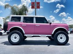 a pink truck parked in a parking lot next to a flag pole and palm trees