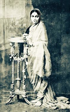 an old photo of a woman standing next to a table with a vase on it