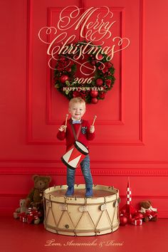 a little boy standing on top of a drum in front of a red wall with christmas decorations