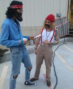 two men with long hair and beards standing next to each other in front of a building