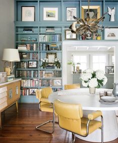 a dining room table with yellow chairs in front of bookshelves and pictures on the wall