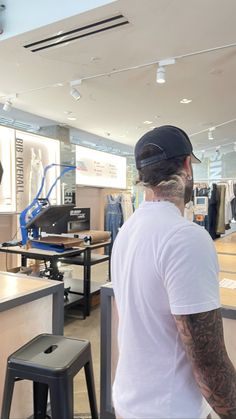 a man standing in front of a store counter with his back to the camera, wearing a white shirt and black hat