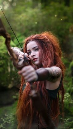 a woman with long red hair holding a bow and arrow in her hands while standing on mossy ground