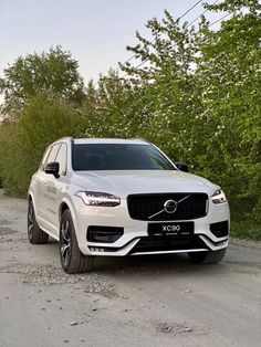 a white volvo suv parked on the side of a dirt road next to some trees