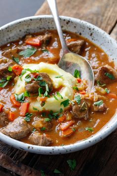 a white bowl filled with meat and vegetable soup on top of a wooden table next to a spoon