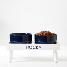 two blue bowls with food in them sitting on a white tray that says rocky and is placed next to each other