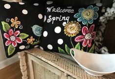 a welcome spring pillow sitting on top of a wooden bench next to a bowl and vase