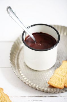 there is a cup of chocolate and some cookies on the plate with one spoon in it